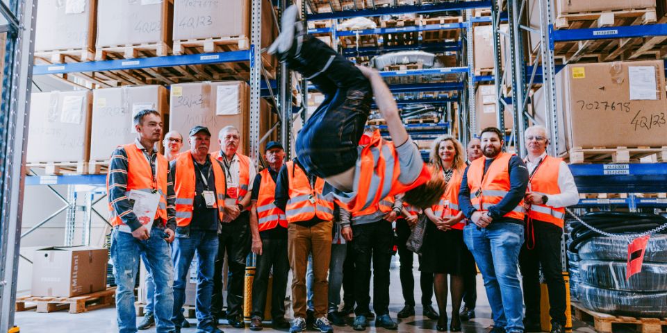 Eine Person macht in einer Lagerhalle einen Rückwärtssalto, während im Hintergrund eine Gruppe von Personen in orangefarbenen Sicherheitswesten steht. Sie sind von Regalen umgeben, die mit großen Kisten und diversem technischen Sortiment gefüllt sind.